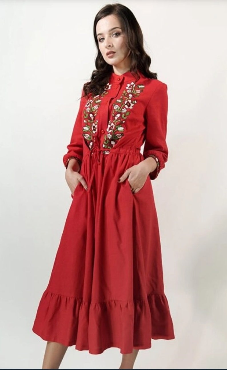May include: A red dress with long sleeves and a ruffled hem. The dress features floral embroidery around the collar and down the front. It has a drawstring waist and pockets.
