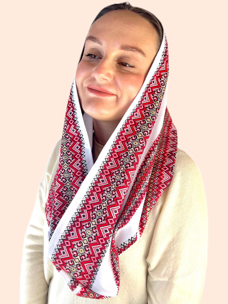 May include: A white headscarf with red and black embroidered geometric patterns. The scarf is folded and worn around the head.