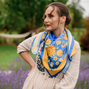 May include: A woman wearing a white dress with a floral pattern and a blue and yellow patterned scarf with sunflowers. She is standing in a field of lavender.