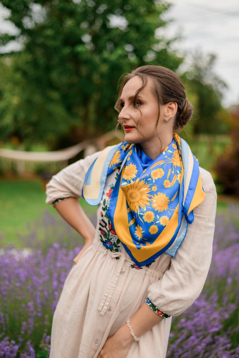 May include: A woman wearing a white dress with a floral pattern and a blue and yellow patterned scarf with sunflowers. She is standing in a field of lavender.