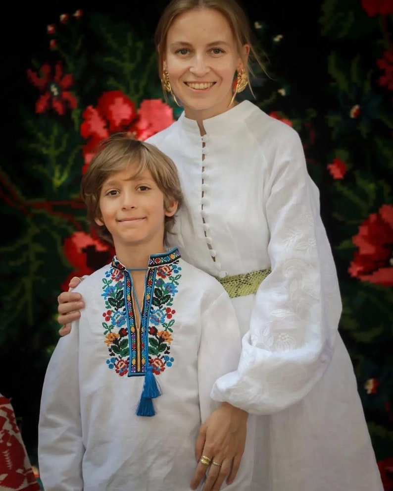 May include: A woman and a young boy wearing traditional Ukrainian embroidered shirts, or vyshyvankas. The woman is wearing a white linen shirt with a green belt and gold earrings. The boy is wearing a white shirt with a colorful floral embroidery pattern and a blue tassel. They are standing in front of a floral patterned background.