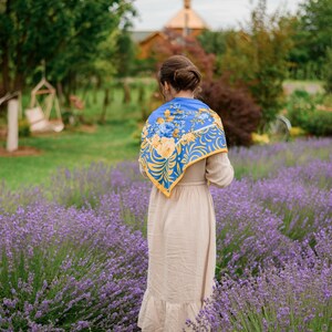 May include: A woman wearing a beige dress and a blue and yellow floral scarf stands in a field of lavender.