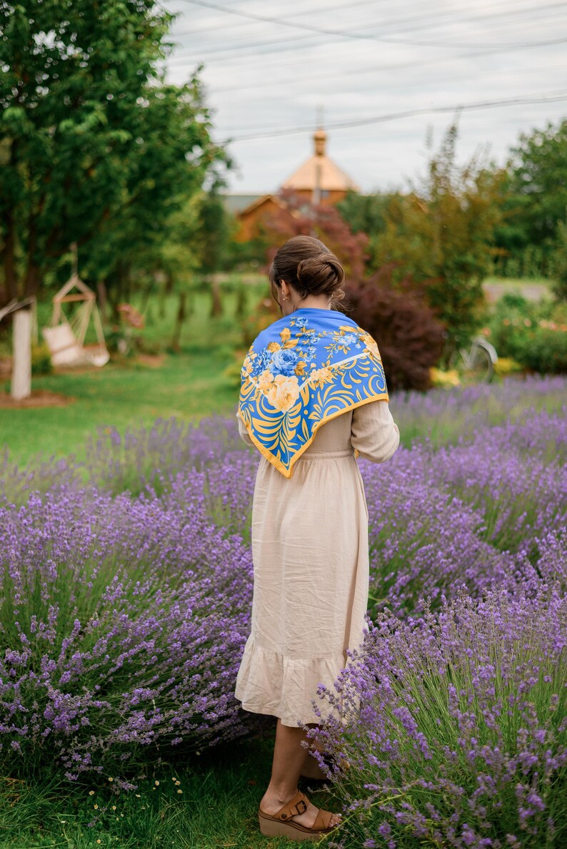 May include: A woman wearing a beige dress and a blue and yellow floral scarf stands in a field of lavender.