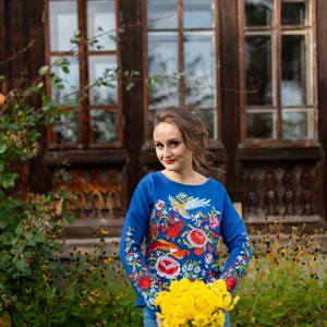 Puede incluir: Una mujer con una camisa azul bordada con un patr&oacute;n floral sostiene un ramo de flores amarillas frente a un edificio de madera con ventanas.