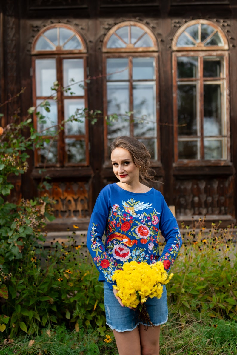 Puede incluir: Una mujer con una camisa azul bordada con un patr&oacute;n floral sostiene un ramo de flores amarillas frente a un edificio de madera con ventanas.
