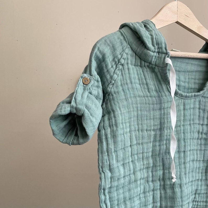 May include: A light green hooded baby shirt with a button on the sleeve. The shirt is made of a soft, textured fabric.