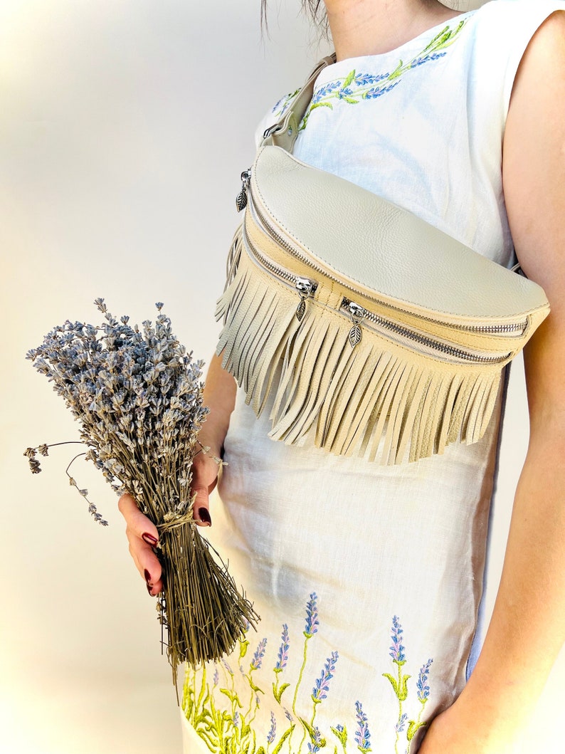 Puede incluir: Un cintur&oacute;n de cuero color crema con flecos y cremalleras plateadas. La bolsa se lleva sobre un vestido blanco con bordados de lavanda. Un manojo de lavanda seca se sostiene en la mano.