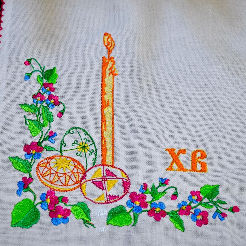 Puede incluir: Toalla de cocina de lino blanco bordada con una vela amarilla, tres huevos de Pascua coloridos y ramitas de flores verdes. Las letras cir&iacute;licas "XB" est&aacute;n bordadas en naranja en la esquina superior derecha.