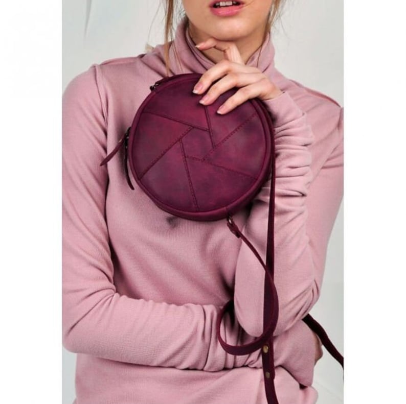 May include: A round, burgundy-colored crossbody bag with a geometric design. The bag features a long, thin strap and a zipper closure. The bag is made of a textured material and is held by a person wearing a pink turtleneck.