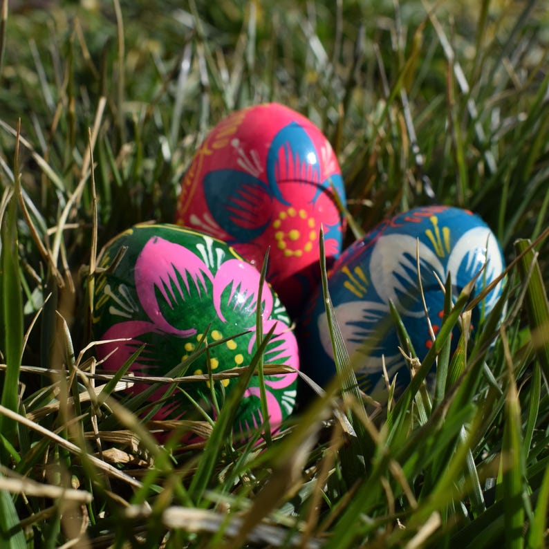 May include: Three decorated Easter eggs with floral designs in a bed of green grass. The eggs are painted in bright colors, including pink, blue, and green. The floral patterns are detailed and add a festive touch.