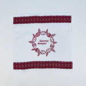 May include: White Easter towel with red embroidered borders and a central wreath design. The wreath features decorated Easter eggs and the text "Christ is Risen!" in Cyrillic script. A decorative kitchen or bathroom accessory.