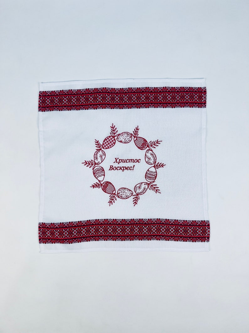 May include: White Easter towel with red embroidered borders and a central wreath design. The wreath features decorated Easter eggs and the text "Christ is Risen!" in Cyrillic script. A decorative kitchen or bathroom accessory.