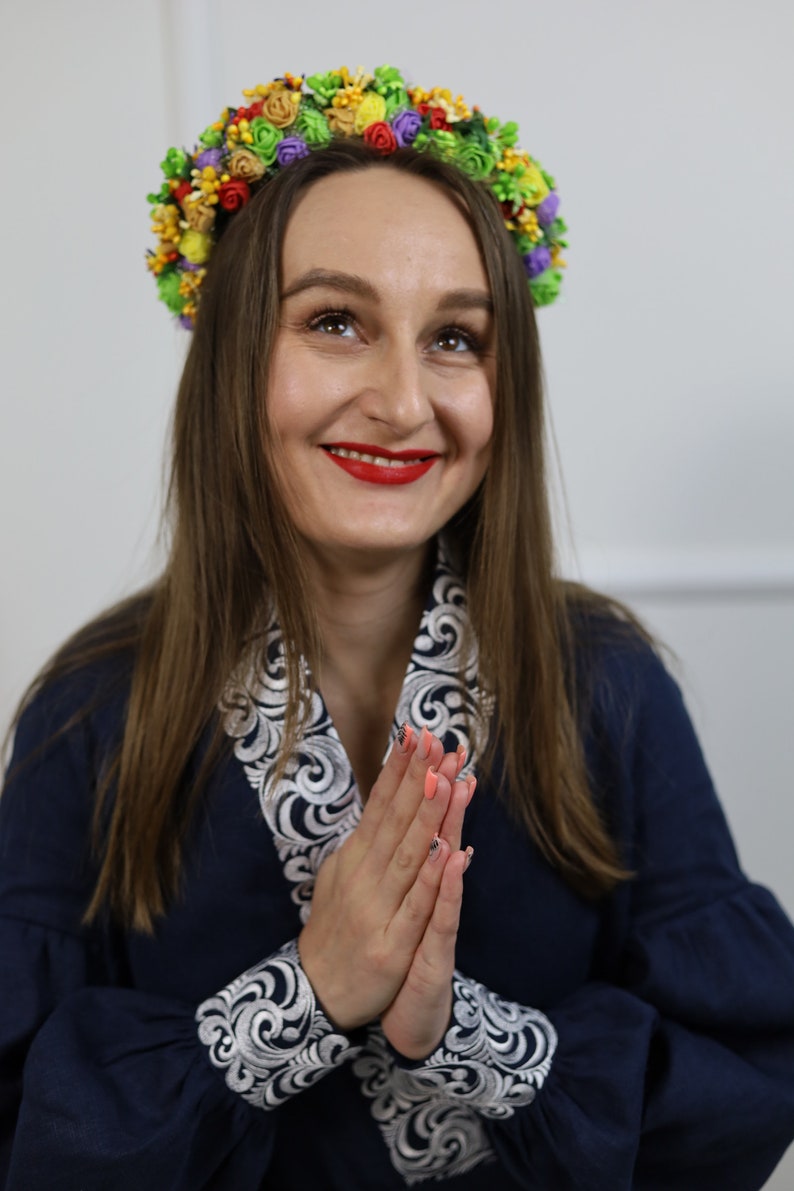 K&ouml;nnte beinhalten: Eine Frau tr&auml;gt einen farbenfrohen Blumenkranz und ein marineblaues Kleid mit wei&szlig;-silberner Stickerei. Der Kranz zeigt eine Vielzahl kleiner, bunter Blumen. Die Frau l&auml;chelt und blickt nach oben.