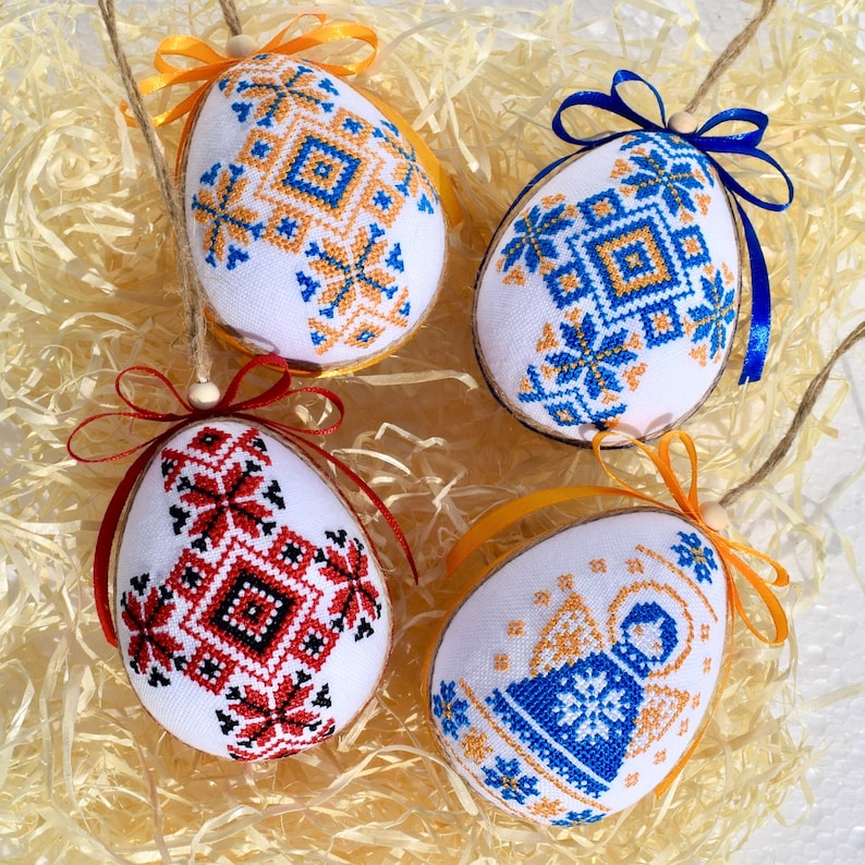 May include: Four white Easter egg ornaments with embroidered folk art designs in blue, orange, and red. Each egg has a ribbon bow and a twine loop for hanging. The eggs are displayed on a bed of straw.