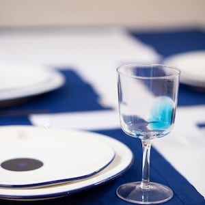 May include: A clear wine glass with a blue liquid inside sits on a blue tablecloth. White plates with blue trim and a black circle are in the background. A knife and a white napkin are also on the table.