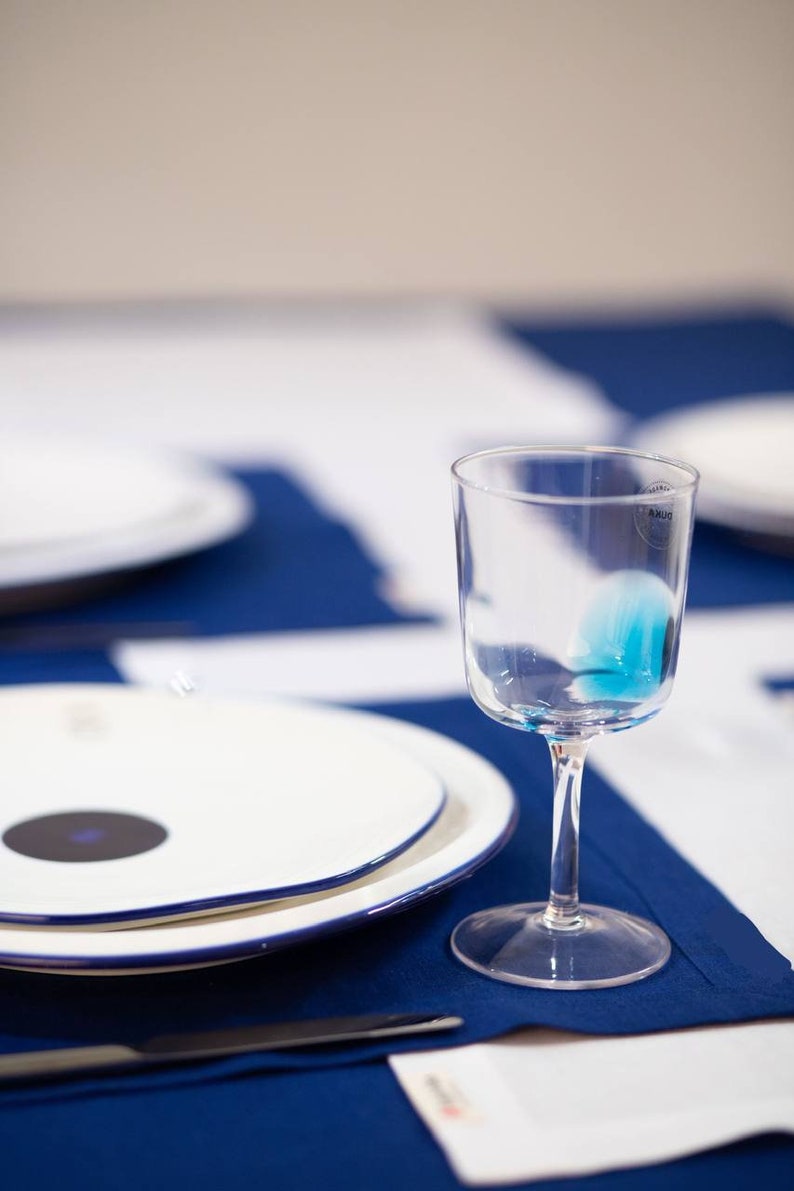 May include: A clear wine glass with a blue liquid inside sits on a blue tablecloth. White plates with blue trim and a black circle are in the background. A knife and a white napkin are also on the table.