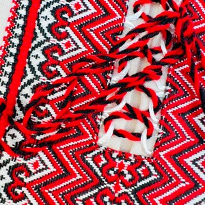 Op de afbeelding: Close-up van een textiel met gedetailleerd borduurwerk. Het ontwerp toont een herhalend geometrisch patroon in rood, zwart en wit. De randen zijn afgewerkt met een rode rand en zwarte en rode kwasten.
