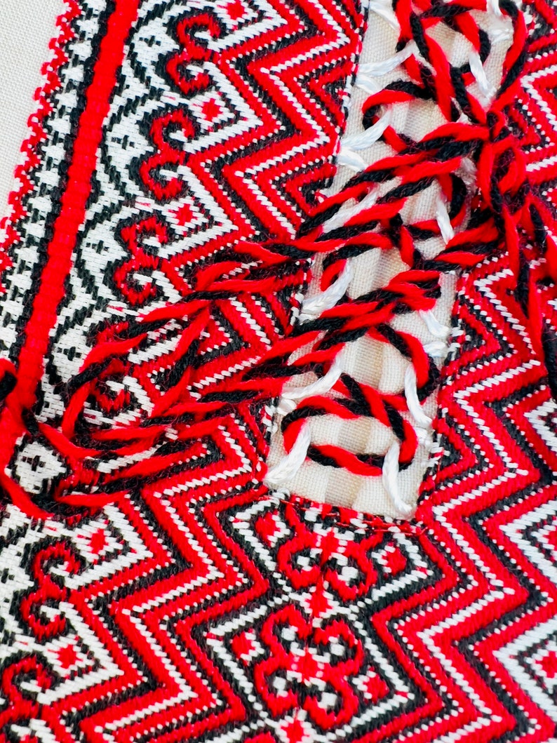 Op de afbeelding: Close-up van een textiel met gedetailleerd borduurwerk. Het ontwerp toont een herhalend geometrisch patroon in rood, zwart en wit. De randen zijn afgewerkt met een rode rand en zwarte en rode kwasten.