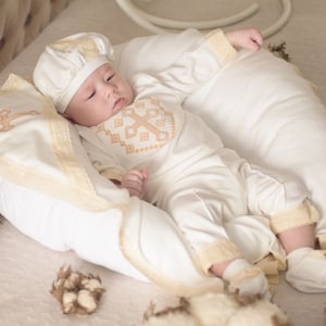 May include: A white baby outfit with a cross embroidered on the chest, a white hat with lace trim, and a white blanket with lace trim. The baby is lying on a white pillow.