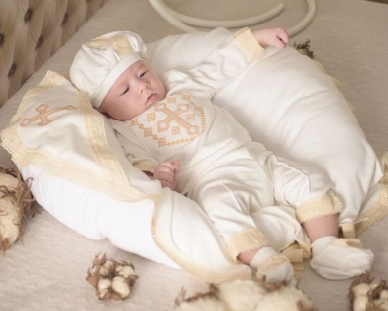 May include: A white baby outfit with a cross embroidered on the chest, a white hat with lace trim, and a white blanket with lace trim. The baby is lying on a white pillow.