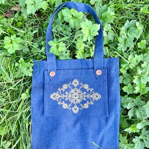 Op de afbeelding: Een blauwe denim tote bag met een gouden geborduurd bloemenmotief. De tas heeft twee bruine knoopsluitingen.