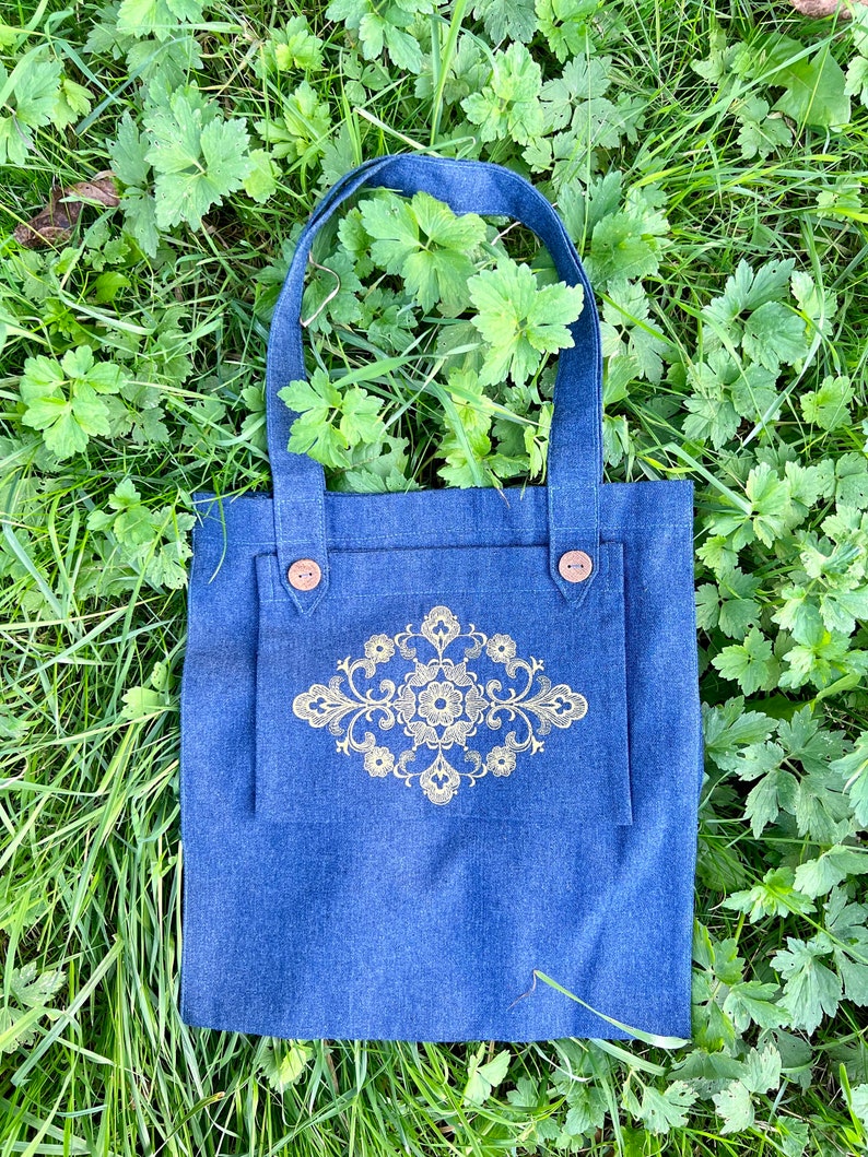 Op de afbeelding: Een blauwe denim tote bag met een gouden geborduurd bloemenmotief. De tas heeft twee bruine knoopsluitingen.