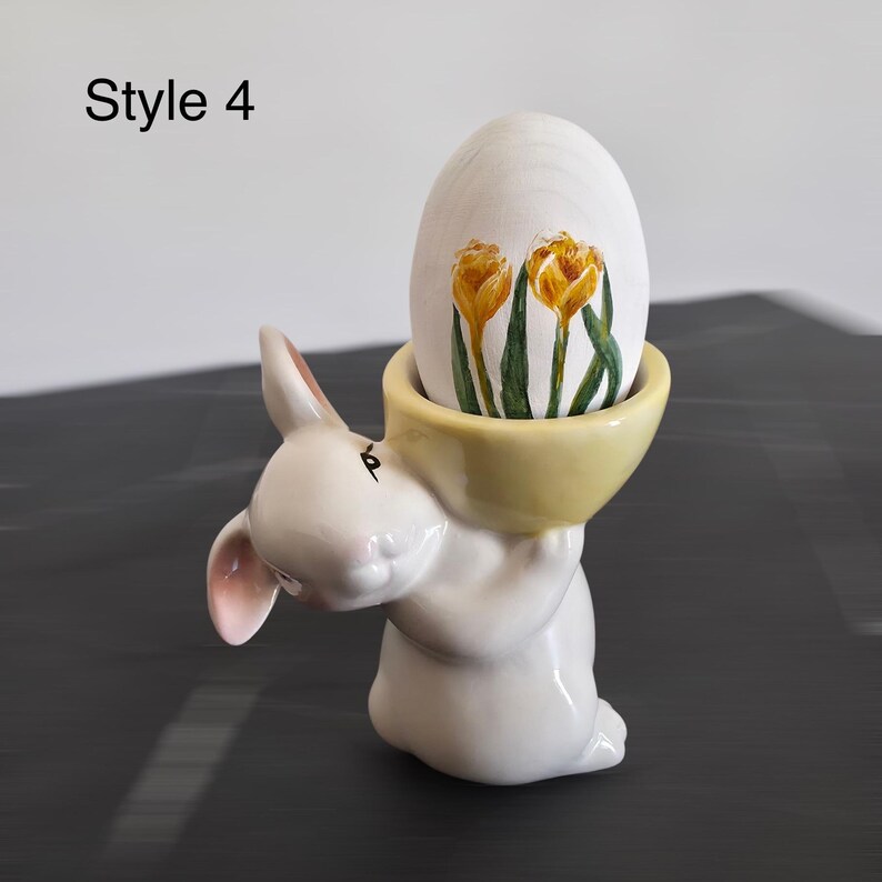 May include: A white ceramic bunny figurine holding a yellow bowl with a wooden egg. The egg is decorated with yellow and green floral artwork. The text "Style 4" is in the upper left corner. The bunny has pink inner ears.