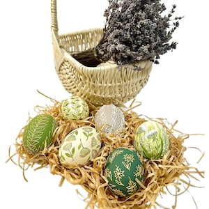 May include: Six hand-painted Easter eggs in a wicker basket with lavender. The eggs are decorated with green, white and gold floral patterns.
