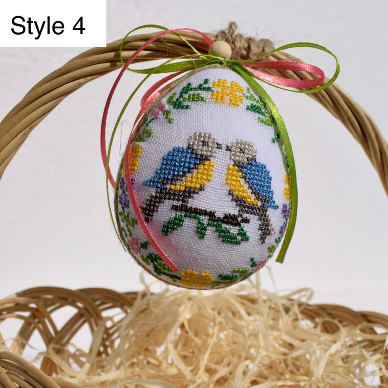 Op de afbeelding: Een wit, eivormig ornament met kruissteek blauwe en gele vogels op een tak, omringd door bloemenborduurwerk. Het ornament hangt aan een gevlochten mand met groene en roze linten. De tekst "Style 4" is zichtbaar.