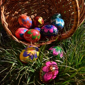 May include: A woven basket overflowing with vibrantly painted Easter eggs. The eggs feature intricate floral designs in shades of red, blue, pink, yellow, and green. The basket sits in a bed of green grass, illuminated by sunlight.
