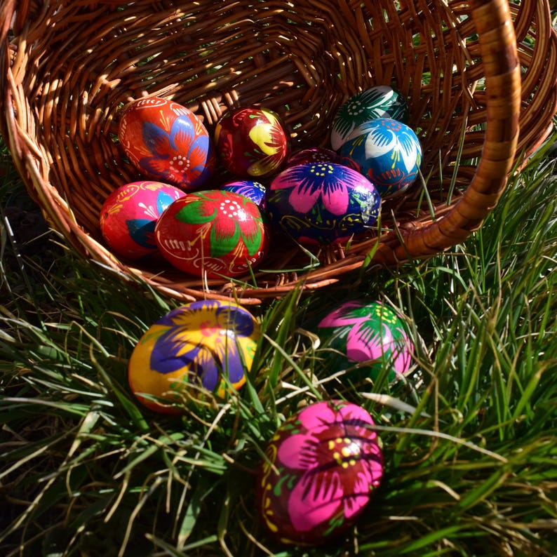 May include: A woven basket overflowing with vibrantly painted Easter eggs. The eggs feature intricate floral designs in shades of red, blue, pink, yellow, and green. The basket sits in a bed of green grass, illuminated by sunlight.