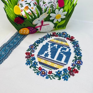 May include: An Easter-themed display featuring a felt basket with faux grass, decorative eggs, and a white bunny. A white tablecloth has a blue and white embroidered border and a central embroidered egg design with blue lettering, surrounded by a floral wreath.