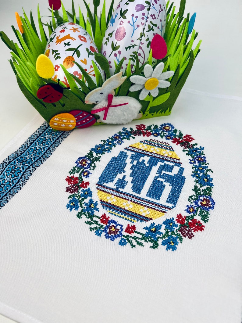 May include: An Easter-themed display featuring a felt basket with faux grass, decorative eggs, and a white bunny. A white tablecloth has a blue and white embroidered border and a central embroidered egg design with blue lettering, surrounded by a floral wreath.