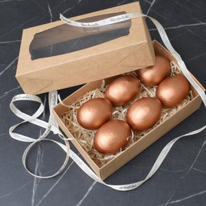May include: A cardboard box with a clear window holds six metallic copper-coloured eggs in straw. A matching lid is open beside it. A white ribbon with "Handmade" text is wrapped around the box.