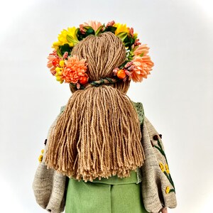 Puede incluir: Mu&ntilde;eca hecha a mano vista desde atr&aacute;s, con un vestido verde y una blusa bordada beige. La mu&ntilde;eca tiene pelo largo de hilo marr&oacute;n y una corona floral con flores amarillas y melocot&oacute;n y bayas naranjas.