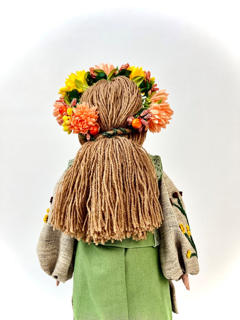 Puede incluir: Mu&ntilde;eca hecha a mano vista desde atr&aacute;s, con un vestido verde y una blusa bordada beige. La mu&ntilde;eca tiene pelo largo de hilo marr&oacute;n y una corona floral con flores amarillas y melocot&oacute;n y bayas naranjas.