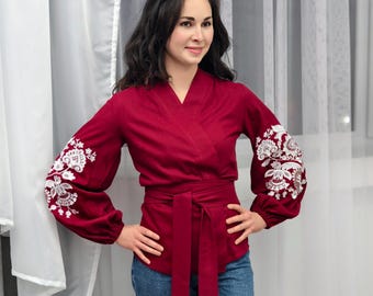 Könnte beinhalten: Eine Frau trägt ein rotes Wickeltop mit weißen, gestickten Blumenmustern an den Ärmeln. Das Oberteil hat einen Bindegürtel in der Taille und die Frau trägt eine blaue Jeans.