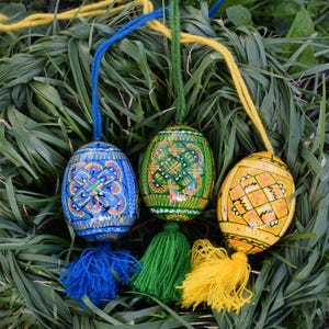 May include: Three vibrantly painted Easter eggs with intricate designs and tassels. The eggs are blue, green, and yellow, each with a unique pattern. They hang against a backdrop of green grass, showcasing the detailed artwork and festive colors.