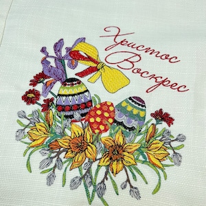 Op de afbeelding: Geborduurde keukendoek met een bloemmotief met gele narcissen, paarse irissen en rode bloemen. Drie kleurrijke paaseieren zijn omringd door de bloemen. De tekst "Христос Воскрес" is geborduurd in rode draad.