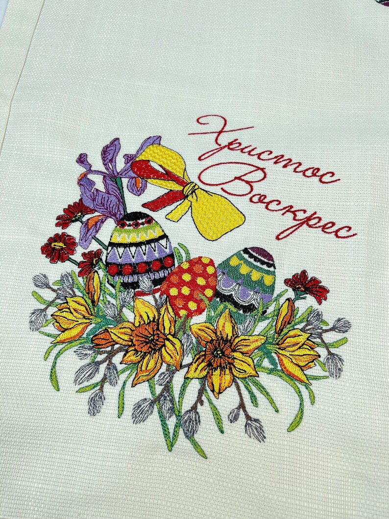 Op de afbeelding: Geborduurde keukendoek met een bloemmotief met gele narcissen, paarse irissen en rode bloemen. Drie kleurrijke paaseieren zijn omringd door de bloemen. De tekst "Христос Воскрес" is geborduurd in rode draad.