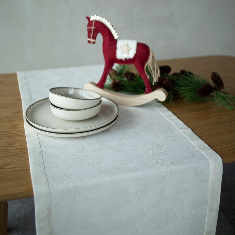 Op de afbeelding: Een witte tafelloper met een subtiel reli&euml;f bloemmotief, gecombineerd met een set cr&egrave;mekleurige borden en een kom. Een rood schommelpaard en dennentakken voegen een feestelijk tintje toe. De tafelloper is ongeveer 30 cm breed.