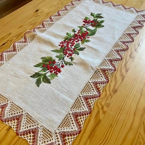 May include: A white linen table runner with red embroidered berries and leaves. The runner has a delicate lace border with red accents.