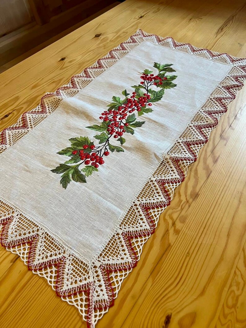 May include: A white linen table runner with red embroidered berries and leaves. The runner has a delicate lace border with red accents.