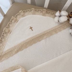 May include: A white hooded towel with a beige lace trim and a small gold cross embellishment. The towel is folded and displayed on a white crib.