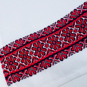 May include: White fabric with a red and black embroidered border. The embroidery features a geometric pattern of interlocking shapes.