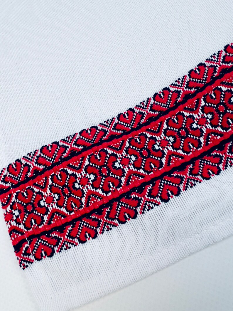 May include: White fabric with a red and black embroidered border. The embroidery features a geometric pattern of interlocking shapes.