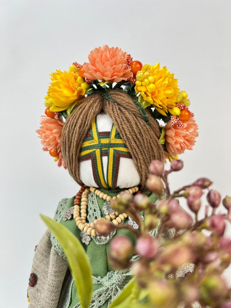 Puede incluir: Mu&ntilde;eca hecha a mano con una corona floral. La mu&ntilde;eca tiene cabello de hilo marr&oacute;n y una cara blanca con detalles bordados. Viste un vestido verde, un chal beige y un collar de cuentas. La corona presenta flores naranjas y amarillas.