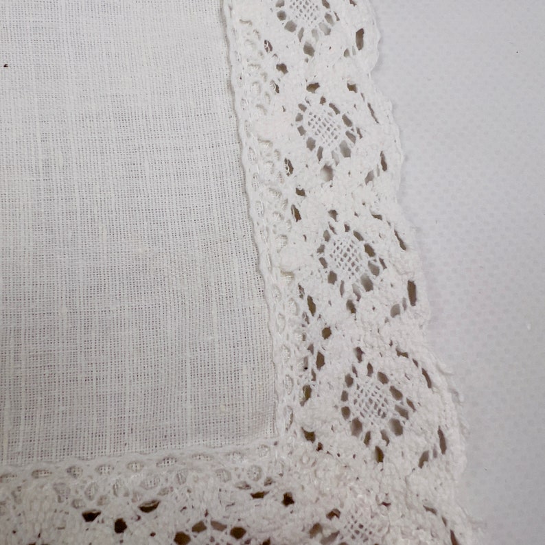 Pu&ograve; includere: Primo piano di un quadrato di lino bianco con un delicato bordo in pizzo traforato. Il pizzo presenta un motivo geometrico ripetuto, che aggiunge un tocco di eleganza al tessuto semplice e strutturato. L'estetica generale &egrave; pulita e classica.