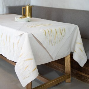 Peut inclure: Une nappe en lin blanc avec des &eacute;pis de bl&eacute; brod&eacute;s en or et beige. La nappe est drap&eacute;e sur une table en bois. Une cafeti&egrave;re dor&eacute;e et une pile d'assiettes blanches sont visibles. Le design est &eacute;l&eacute;gant et rustique.