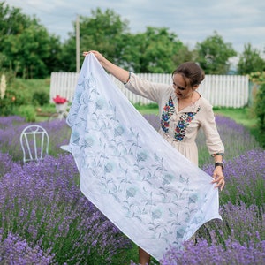Puede incluir: Una bufanda blanca con un dise&ntilde;o floral azul se muestra en un campo de lavanda. La bufanda es rectangular y parece estar hecha de una tela ligera. El fondo muestra un campo de lavanda p&uacute;rpura y una valla blanca.